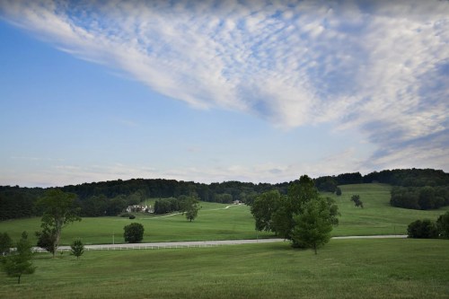 A serene landscape featuring rolling green hills, trees, and a cloudy blue sky. A road runs through the foreground.