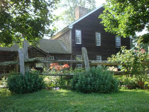 A rustic wooden house surrounded by greenery and a wooden fence, with flowers blooming in the garden.