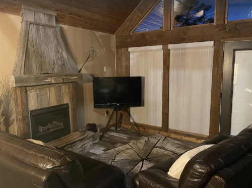 Cozy living room with a wooden fireplace, TV on a stand, and leather sofas, featuring large windows and rustic decor.