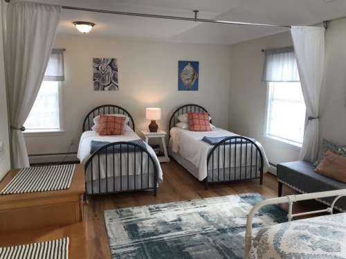 A cozy bedroom with two twin beds, a nightstand, and a seating area, featuring light curtains and decorative artwork.