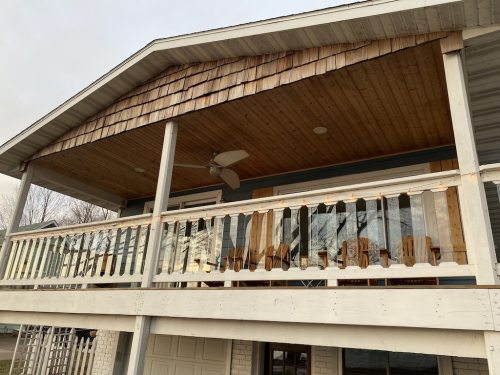 A wooden porch with a shingled roof, featuring a railing and several chairs, under a cloudy sky.