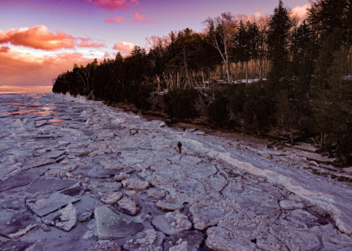 A person walks on a frozen shoreline with ice chunks, surrounded by trees and a colorful sunset sky.