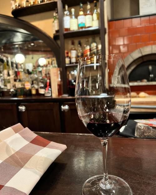 A glass of red wine sits on a bar counter, with shelves of liquor bottles in the background.