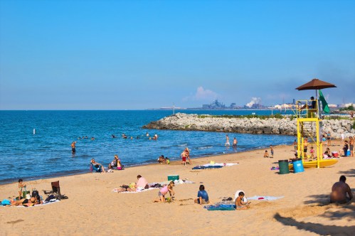 A sunny beach scene with people sunbathing, swimming, and a lifeguard tower by the water's edge.