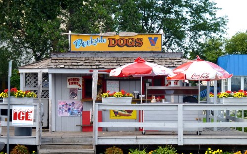 A small food stand named "Dockside Dogs" with red umbrellas, flowers, and a sign advertising hot dogs and ice.