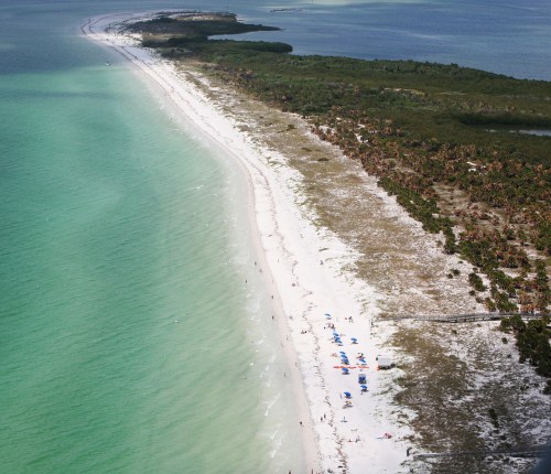 Aerial view of a sandy beach with turquoise waters, lined with palm trees and beachgoers under blue skies.