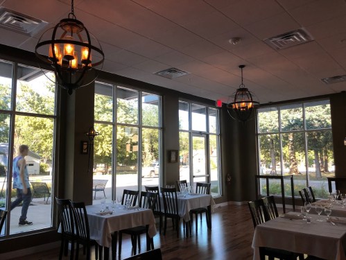 A cozy restaurant interior with tables set for dining, large windows, and a person walking outside.
