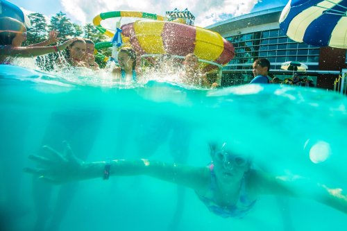 A group of children playing in a pool, with colorful water slides and umbrellas in the background.