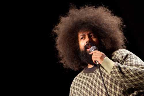 A person with a large afro hairstyle holds a microphone, speaking passionately on stage.