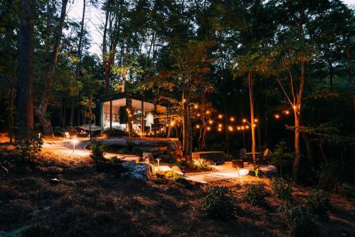 A modern glass cabin surrounded by trees, illuminated by string lights at dusk, creating a cozy outdoor atmosphere.