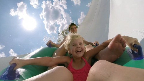 A child and an adult enjoy a water slide, laughing as they descend under a bright sky with fluffy clouds.