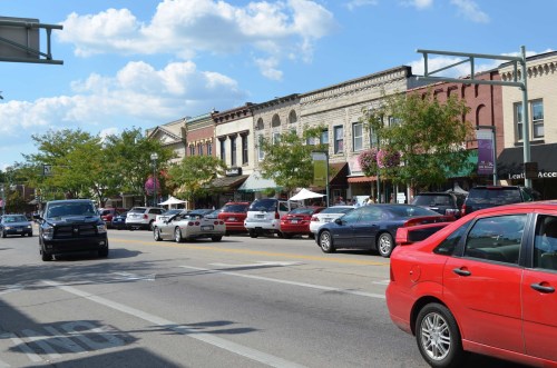 A busy street lined with shops and parked cars, featuring trees and colorful flower baskets under a blue sky.