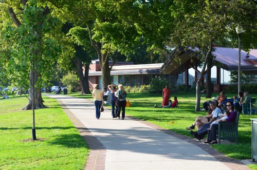 A sunny park scene with people walking on a path, sitting on benches, and enjoying the greenery.