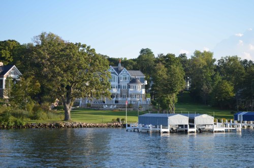 A large, elegant house with a manicured lawn beside a calm lake, featuring a dock with boats. Trees surround the area.