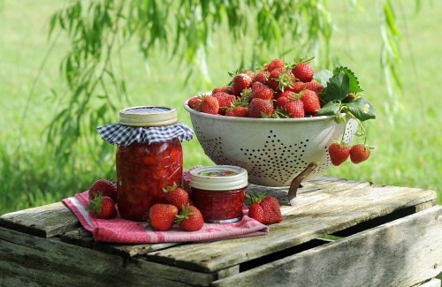 A bowl of fresh strawberries sits on a wooden crate, alongside jars of homemade strawberry jam.