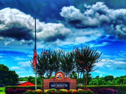 Welcome sign for Bay Minette, Alabama, with a flag at half-mast and vibrant flowers under a cloudy sky.
