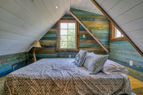 Cozy attic bedroom with a patterned bedspread, decorative pillows, and colorful wooden walls. Natural light from a window.