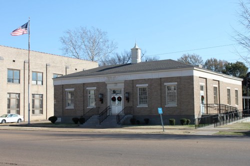A brick building with a peaked roof, featuring a front staircase and American flag, set against a clear blue sky.