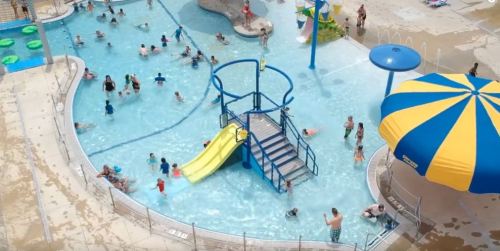 Aerial view of a busy water park with a pool, slides, and colorful umbrellas, filled with families enjoying the water.