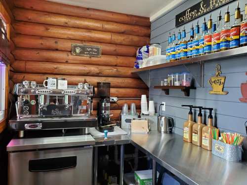 A cozy coffee shop corner featuring an espresso machine, syrups, cups, and a wooden wall.