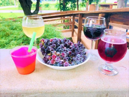 A table with a bowl of grapes and four glasses of colorful drinks, set outdoors on a sunny day.