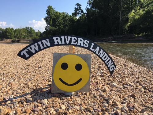 A sign reading "Twin Rivers Landing" with a smiling face, set on a rocky riverbank surrounded by trees.