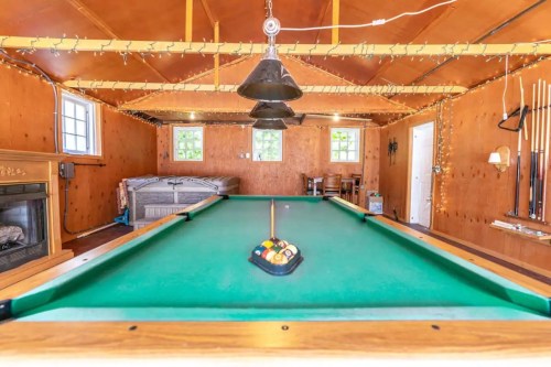 A cozy game room featuring a green pool table, fireplace, and warm wooden walls, with string lights adding ambiance.