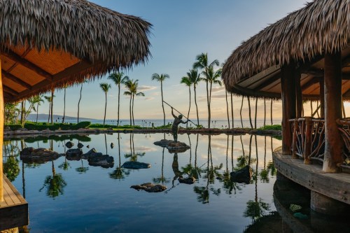 A serene tropical scene with a fisherman standing on rocks, surrounded by palm trees and reflecting water at sunset.