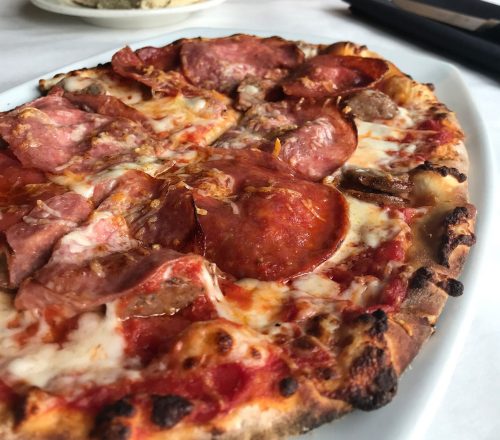 A close-up of a pepperoni and sausage pizza on a white plate, with a loaf of bread and salt shakers in the background.