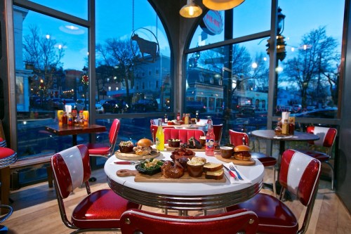 A cozy diner table set with various burgers and sides, with a view of a street outside at dusk.