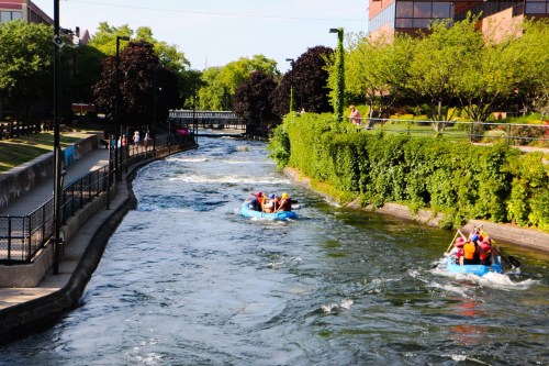 A group of people in blue rafts navigating a river surrounded by greenery and buildings on a sunny day.