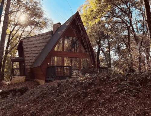 A cozy A-frame cabin nestled in a wooded area, surrounded by trees and sunlight filtering through the leaves.