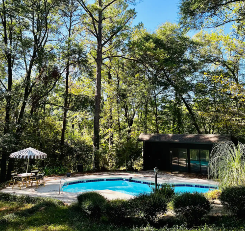 A serene backyard pool surrounded by trees, with a patio, umbrella, and a small building in the background.