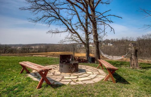 A serene outdoor scene featuring a fire pit surrounded by stone, with benches and a hammock under a tree.