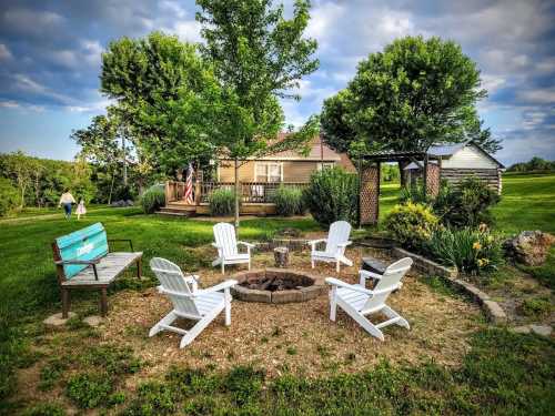 A cozy outdoor fire pit area with white chairs, surrounded by greenery and a house in the background.