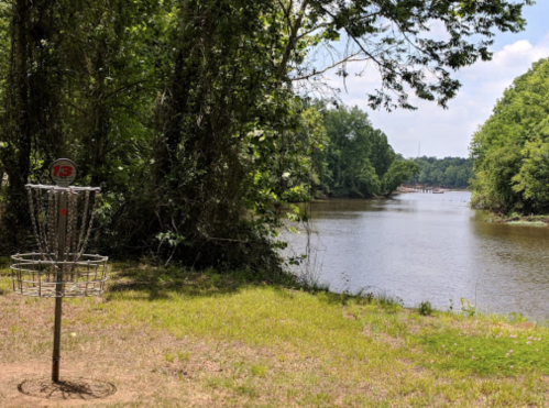 A disc golf basket near a river, surrounded by lush greenery and trees under a clear blue sky.