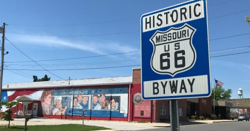 Sign for Historic Route 66 in front of a colorful mural on a building, with power lines and trees in the background.