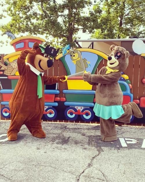 Two cartoon bear mascots in costumes playfully sword fighting with foam swords in front of a colorful train backdrop.