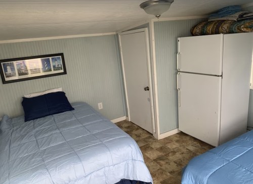 A small room with two beds, a refrigerator, and a door, featuring light blue walls and a framed picture.