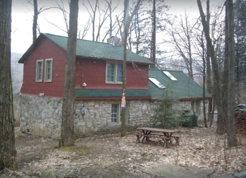 A rustic cabin with a stone base and red wooden upper, surrounded by trees and a picnic table in a wooded area.