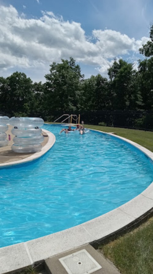 A curved swimming pool with clear blue water, surrounded by green trees and inflatable floats.