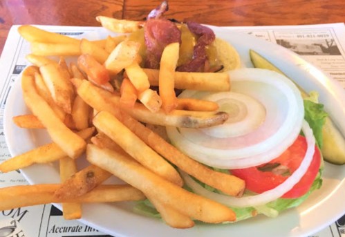 A plate of fries with a side of lettuce, tomato, onion, pickles, and a burger topped with bacon and mustard.