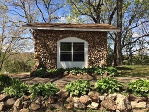 A small stone cottage with a white window, surrounded by greenery and trees under a clear blue sky.