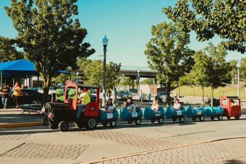 A small train with blue cars and passengers rides through a park on a sunny day, surrounded by trees and buildings.
