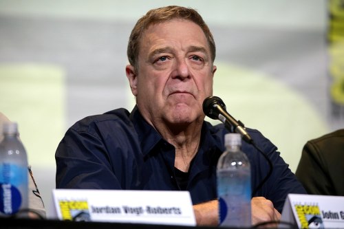 A middle-aged man with short hair sits at a panel, looking serious, with a water bottle in front of him.