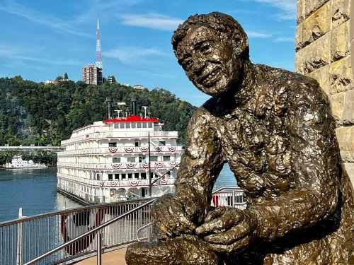 A bronze statue of a seated man overlooks a riverboat on the water, with a city skyline in the background.