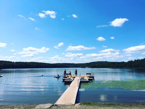 A serene lake scene with a dock, people enjoying the water, and a clear blue sky with fluffy clouds.