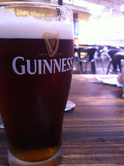 A glass of Guinness beer sits on a wooden table, with a frothy head and a blurred background of a casual setting.