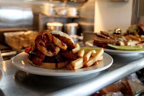 A close-up of a burger topped with onion rings and served with French fries on a white plate in a kitchen setting.