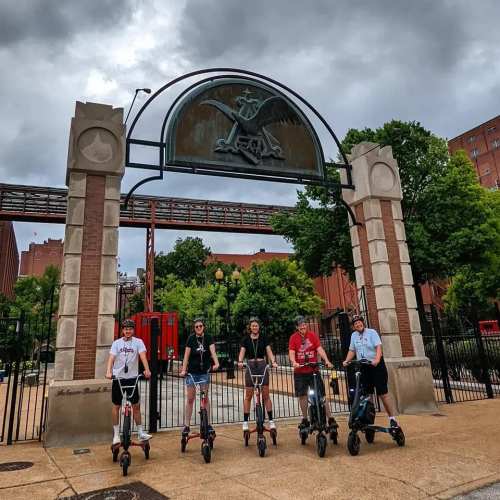 Five people stand on electric scooters in front of a large archway with a decorative emblem, under a cloudy sky.
